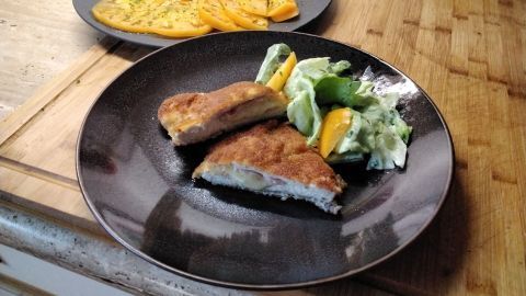 Es liegt ein Teller mit Puten Cordon bleu und einem bunten Salat auf einem Holzbrett; im Hintergrund sieht man einen weiteren Teller.