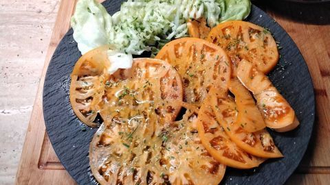 Scheiben von Ananastomaten liegen mit Kräutern auf einer schwarzen Platte, umgeben von frischem Salat und einem Holzbrett.