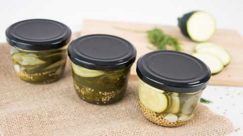 Drei Gläser mit eingelegten Zucchini stehen auf einem Tuch, neben frischen Zucchini und Gewürzen auf einem Holzbrett.