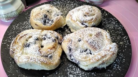 Auf einem schwarzen Teller liegen vier frische Blaubeer-Hefeschnecken, bestäubt mit Puderzucker, auf einem rosa Tisch.