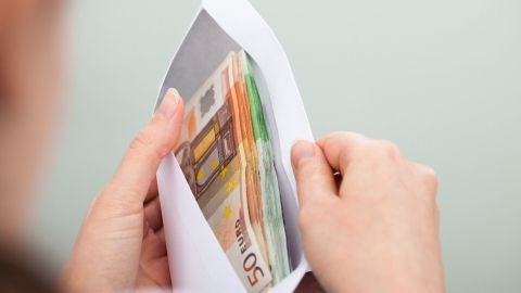 Eine Hand hält einen Umschlag mit Geldscheinen. Im Hintergrund ist eine schlichte, helle Wand zu sehen.