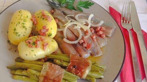Ein Teller zeigt Matjes in Specksoße, umgeben von Kartoffeln und grünem Spargel, angerichtet auf einem karierten Tisch.