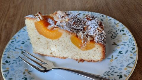 Ein Stück Kuchen mit versunkenen Aprikosen und Mandeln liegt auf einem blümchenmusternden Teller neben einer Gabel.