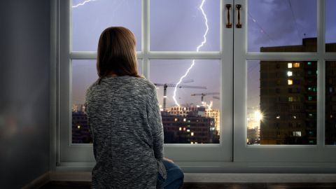 Ein Mädchen sitzt auf dem Boden und schaut aus einem Fenster, während ein Blitz in der Nacht über der Stadt einschlägt.