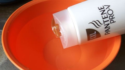 Eine Flasche Pantene Pro-V kippt und gibt eine klare Flüssigkeit in eine orange Schüssel, die auf einer dunklen Oberfläche steht.