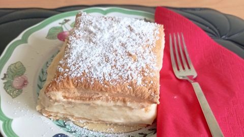 Eine Cremeschnitte aus Blätterteig liegt auf einem dekorativen Teller und wird von einer roten Serviette begleitet.