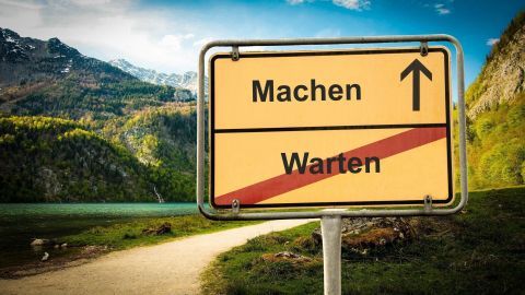 Ein gelbes Verkehrsschild steht am Wegesrand und fordert auf, „Machen“ statt „Warten“ und zeigt die Richtung an.