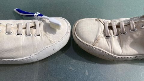 Weiße Sneaker liegen auf einer glatten Fläche, während eine Zahnbürste darauf abgestützt wird, um sie zu reinigen.