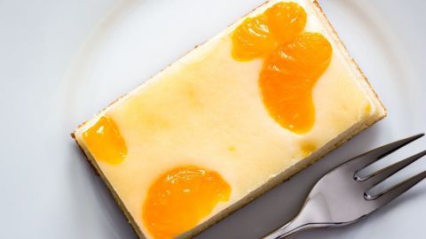 Eine Käsesahnetorte mit Mandarinen liegt auf einem weißen Teller, verziert mit fruchtigen Orangenscheiben und einem Gabel daneben.