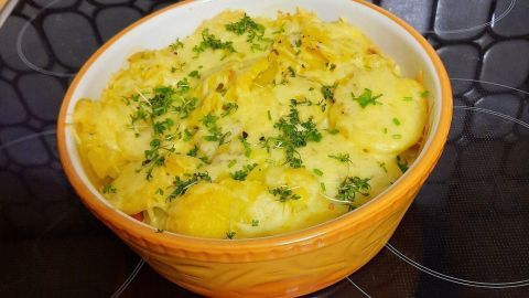 Ein goldbraunes Kartoffelgratin mit grünen Kräutern bäckt in einer orangefarbenen Schüssel auf einem Kochfeld.
