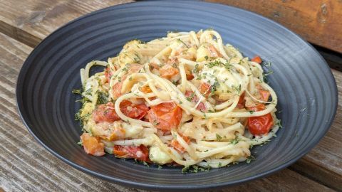 Eine schwarze Schale enthält köstliche Pasta mit Ofengemüse, garniert mit frischen Kräutern, auf rustikalem Holz.