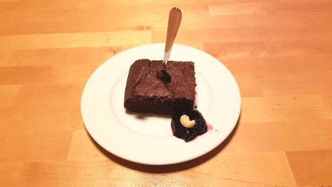 Ein quadratisches Stück Brownie liegt auf einem weißen Teller, begleitet von einem Klecks Beerenkompott und einer Cashew.