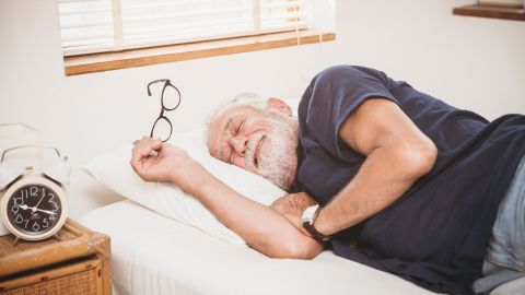 Ein älterer Mann schläft glücklich im Bett, seine Brille liegt neben ihm, während ein Wecker auf dem Nachttisch steht.