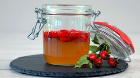 Ein Glas mit goldenem Sauerhonig enthält leuchtend rote Hagebutten, auf einem schwarzen Untergrund platziert.