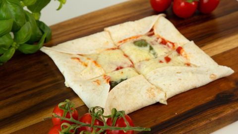 Eine Wrap-Pizza mit Käserand liegt auf einem Holzbrett, umgeben von frischen Tomaten und Basilikum.