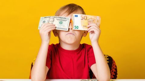 Ein Junge hält je einen Dollar- und Euro-Schein vor sein Gesicht, schaut nachdenklich auf einem gelben Hintergrund.