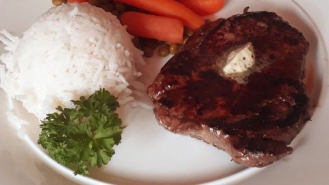 Saftige Rinderlende mit Kräuterbutter liegt neben Reis, Karotten und Erbsen auf einem weißen Teller, serviert auf Holztisch.