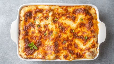Eine goldbraune Lasagne, die mit frischen Kräutern garniert ist, steht in einer weißen Auflaufform auf einer grauen Oberfläche.