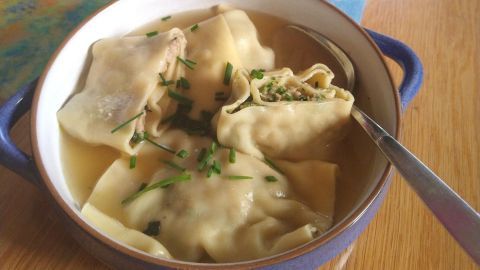 Eine Schüssel mit selbstgemachten Maultaschen in Brühe, garniert mit Schnittlauch, auf einem Holztisch neben einer Blume.