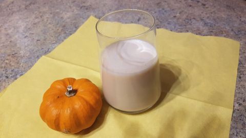 Ein Glas Kürbismilch steht auf einem gelben Tuch, neben einem kleinen orangefarbenen Kürbis in einer gemütlichen Küche.