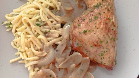 Zarte Hähnchenkeulen liegen in einer cremigen Weißwein-Zwiebelsoße, umgeben von Spätzle auf einem grauen Teller.
