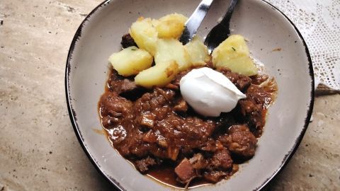 Ein herzhaftes Szegediner Gulasch liegt in einer Schüssel, begleitet von gekochten Kartoffeln und einem Klecks Sauerrahm.