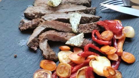 Zarte Scheiben Flanksteak liegen auf einem dunklen Teller, umgeben von buntem Pfannengemüse aus Paprika und Karotten.