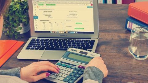 Eine Person nutzt einen Taschenrechner, während sie auf einem Laptop mit Finanzdaten arbeitet, um Spartipps zu entwickeln.
