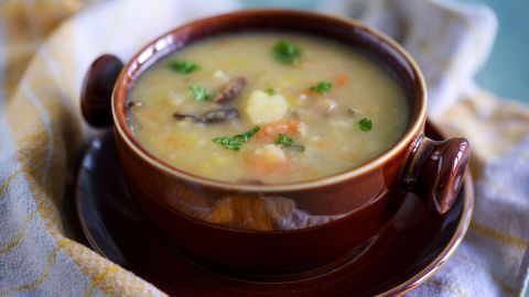 Eine braune Schüssel mit Kartoffel-Gemüse-Suppe steht auf einem Teller, garniert mit frischer Petersilie und umgeben von einem Geschirrtuch.