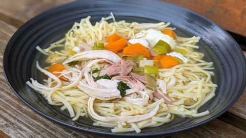 Ein schwarzer Teller mit Hühnersuppe, in dem zarte Hühnerstücke, Nudeln und buntes Gemüse schwimmen.