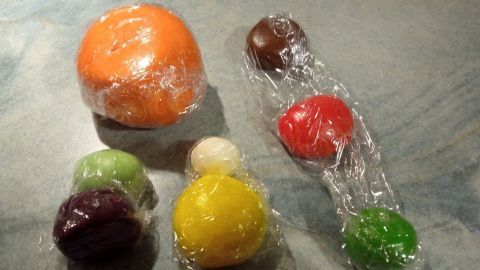Bunte Fondantstücke, eingewickelt in Folie, liegen auf einer Arbeitsfläche und warten auf das Eintönen mit Lebensmittelfarbe.