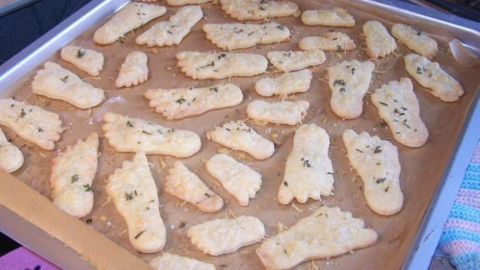 Käsefüße aus Mürbeteig liegen auf einem Blech, frisch gebacken, in Form von kleinen Füßen, mit Kräutern bestreut.