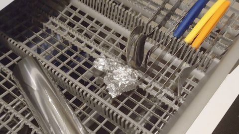 Edelstahlbesteck und eine Aluminiumfolie liegen in einem Geschirrspülerkorb, der für die Reinigung bereit ist.