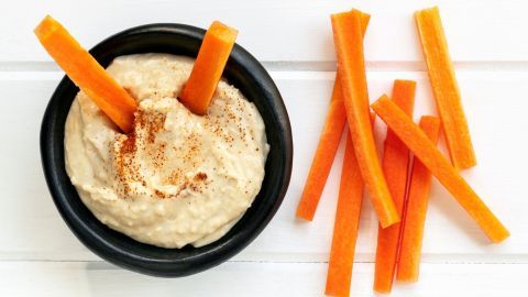 Eine schwarze Schüssel mit Hummus hat Karottensticks, die darin stecken, während weitere Karottensticks daneben liegen.