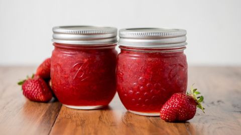 Zwei Gläser mit rot-glänzender Marmelade stehen auf einem Holztablett, umgeben von frischen Erdbeeren.