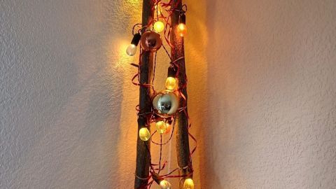 Ein rustikales Dekostück aus zwei Ästen, dekoriert mit bunten Lichtern und Weihnachtskugeln, steht an der Wand.