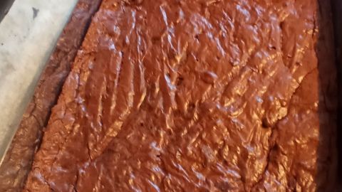 Ein quadratischer Brownie-Kuchen liegt in einer Backform auf Backpapier, bereit zum Servieren oder Genießen.
