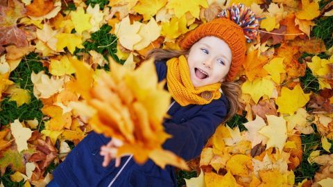 Ein Kind liegt fröhlich auf bunten Herbstblättern und wirft eine Handvoll gelber Blätter in die Luft, umgeben von Natur.