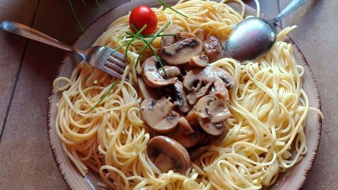 Ein Teller mit Spaghetti wird mit Pilzen garniert, umgeben von roten Tomaten und einem Kräutertopf.