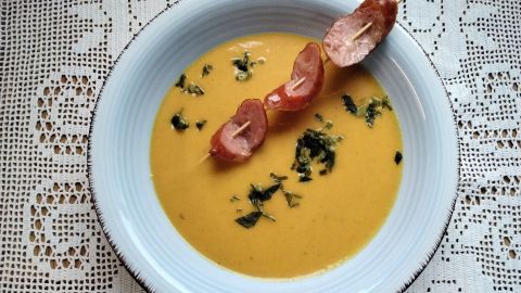 Eine cremige Karottensuppe wird mit Schinkenstücken garniert, serviert in einer hellblauen Schüssel auf einer weißen Tischdecke.