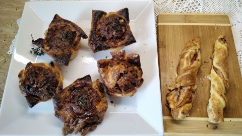 Sechs gefüllte Muffins aus Blätterteig stehen auf einem weißen Teller, neben zwei gedrehten Teigstücken auf einem Holzbrett.