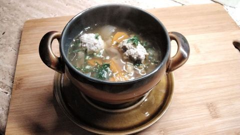 Eine dampfende Schüssel Bratwurstklößchen-Suppe steht auf einem Holzbrett, umgeben von einer gemütlichen Küchenszenerie.