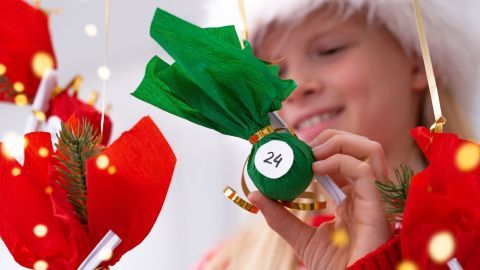 Ein Mädchen mit einem Weihnachtsmann-Hut öffnet einen Adventskalender mit grünen und roten Päckchen, während es lächelt.