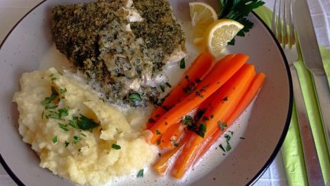 Ein Fischfilet mit Kräuterkuste liegt neben Möhren und Kartoffelpüree auf einem Teller, umgeben von einer weißen Tischdecke.