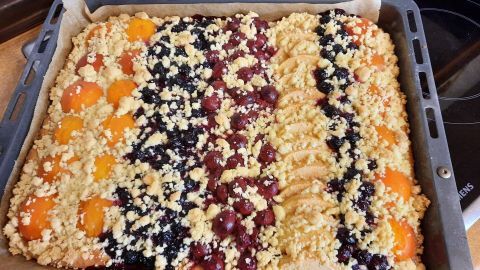 Ein großer, viereckiger Kuchen mit bunten Obstbelägen und Streuseln auf einem Blech, frisch gebacken in einer heimischen Küche.