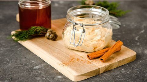 Eine offene Glasbehälter mit Butter steht auf einem Holzbrett, daneben liegen Zimtstangen und ein Glas mit Honig.
