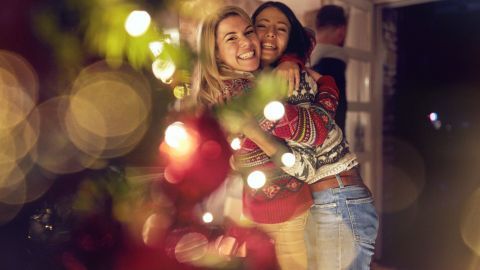 Zwei fröhliche Frauen umarmen sich in festlicher Umgebung, umgeben von Weihnachtslichtern und einer warmen, gemütlichen Atmosphäre.