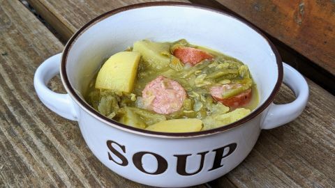 Ein rustikaler Suppentopf mit Kartoffeln und Mettwurst steht auf einer Holzoberfläche, gefüllt mit einer herzhaften Bohnensuppe.