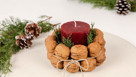 Ein rotes Candle steckt in einem Walnuss-Kerzenhalter, umgeben von frischem Tannengrün und Tannenzapfen auf einem hellen Tisch.