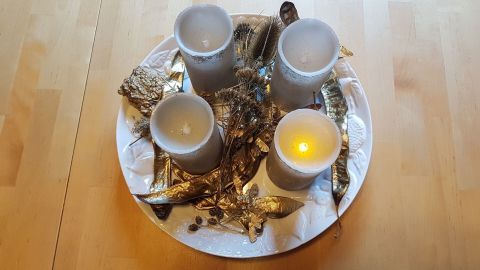 Vier dekorative Kerzen stehen auf einem weißen Teller, umgeben von goldenen Blättern, die eine natürliche Weihnachtsdeko schaffen.